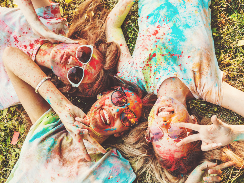 Fototapeta premium Three happy beautiful girls making party at Holi colors festival in summer time.Young smiling women friends having fun after music event at sunset.Models lying on the grass in sunglasses.