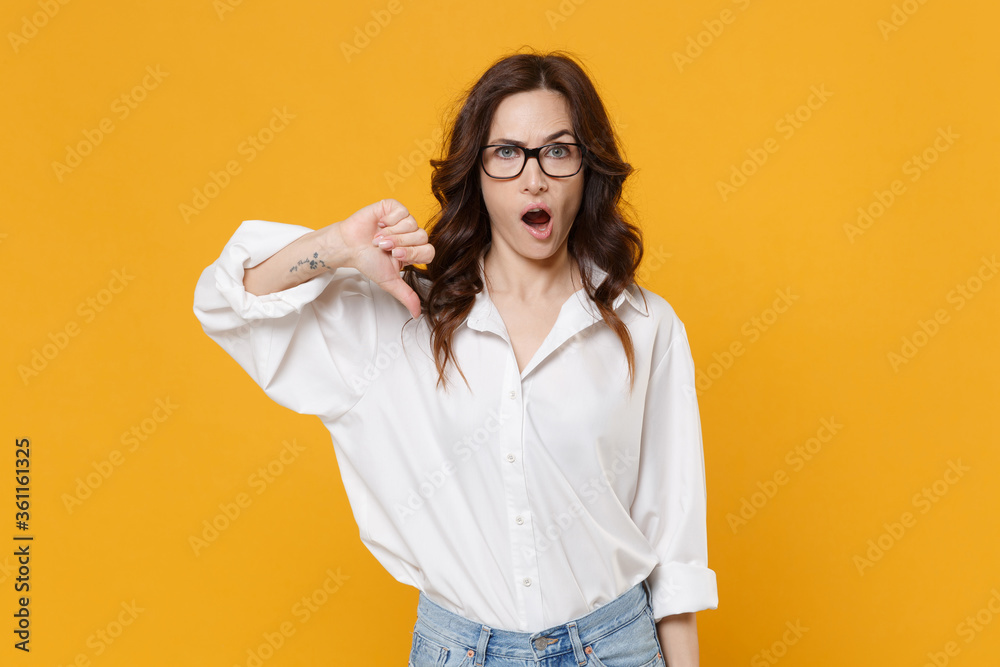 Perplexed young brunette business woman in white shirt glasses isolated on yellow wall background studio portrait. Achievement career wealth business concept. Mock up copy space. Showing thumb down.