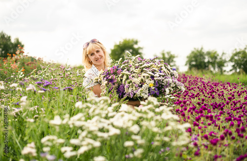 Portrait of a happy woman sitting on a flower bed with a large bouquet of dried flowers in her hands, looking at the camera and smiling. Smiling blonde woman holding flowers grown in the garden