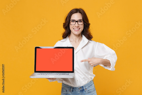 Smiling young business woman in white shirt glasses isolated on yellow background studio. Achievement career wealth business concept. Point index finger on laptop pc computer with blank empty screen.