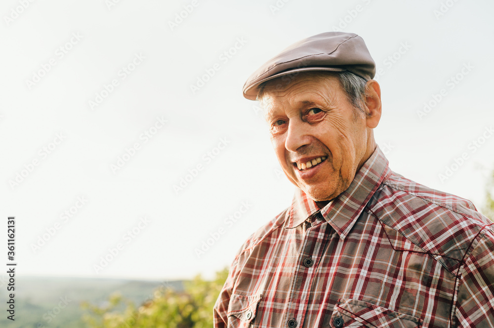 Portrait of old happy man in cap and shirt outdoors at sunset, looking at camera and smiling. Retired man posing at camera, close up photo. Portrait of an old positive grandfather.