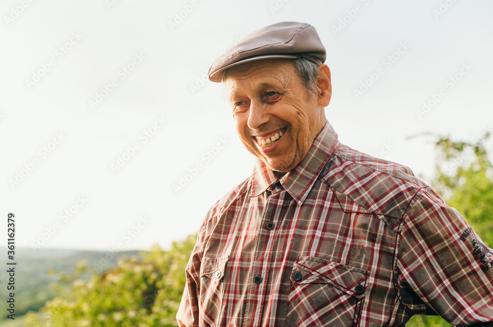Portrait of a smiling old man in a cap and shirt, looking to the side and smiling. Happy retired grandfather, portrait outdoors. Portrait of a retired farmer