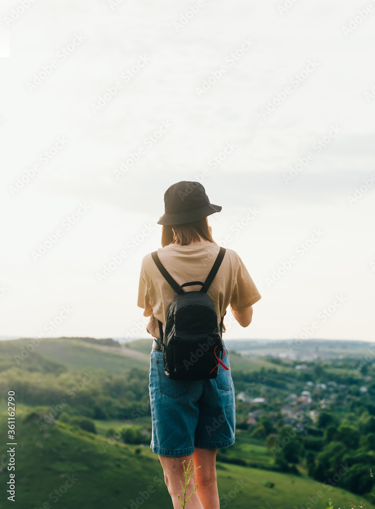 Back of a tourist girl stands on a mountain with a beautiful green landscape. Hipster woman in casual clothes and with a backpack stands on the edge of the mountain.