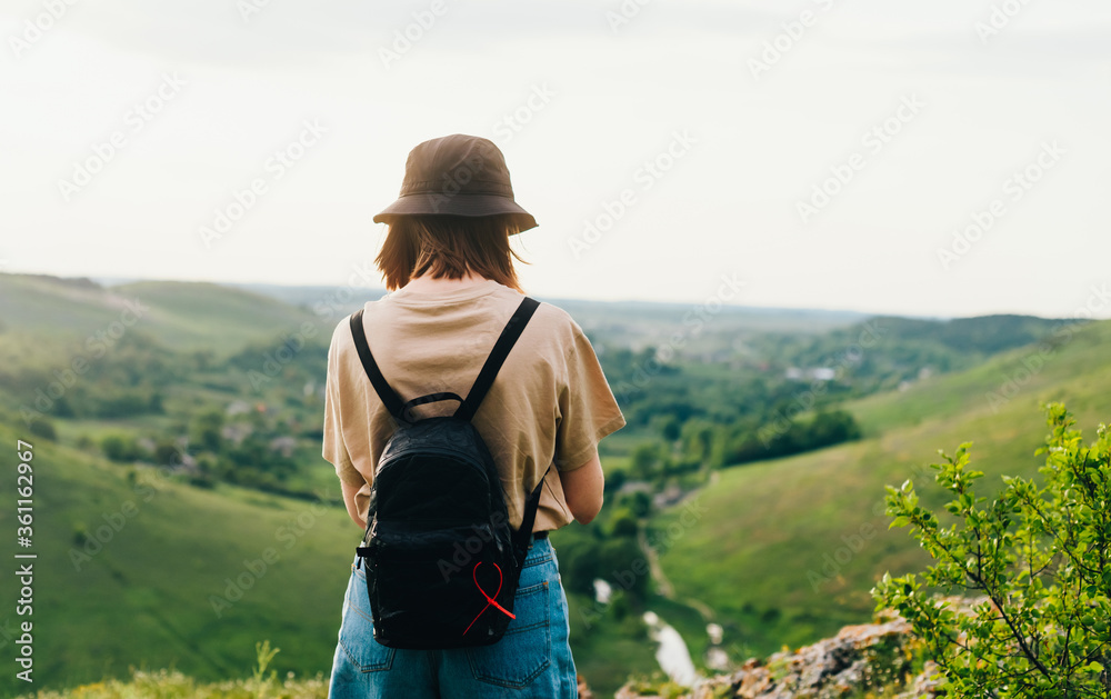 Back of a tourist girl stands on a mountain with a beautiful green landscape. Hipster woman in casual clothes and with a backpack stands on the edge of the mountain. Back of a tourist girl stands on a mountain with a beautiful green landscape. Hipster woman in casual clothes and with a backpack stands on the edge of the mountain.