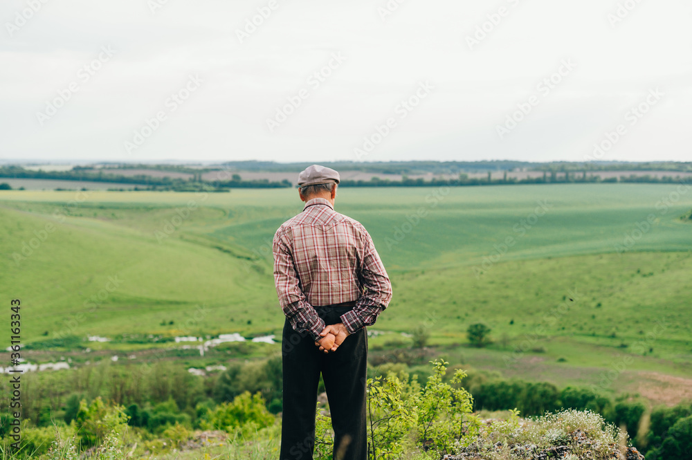 A retired man in a shirt and cap stands on top of a mountain with a beautiful green landscape. Photo of the grandfather of the pensioner from a back on a mountain walk. Pensioner travels