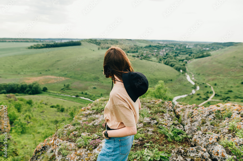 The back of a stylish hipster girl stands on a hill and looks at the beautiful landscape from the top of the mountain, standing on a rock. Woman tourist enjoys the landscape from the mountain Tovtry