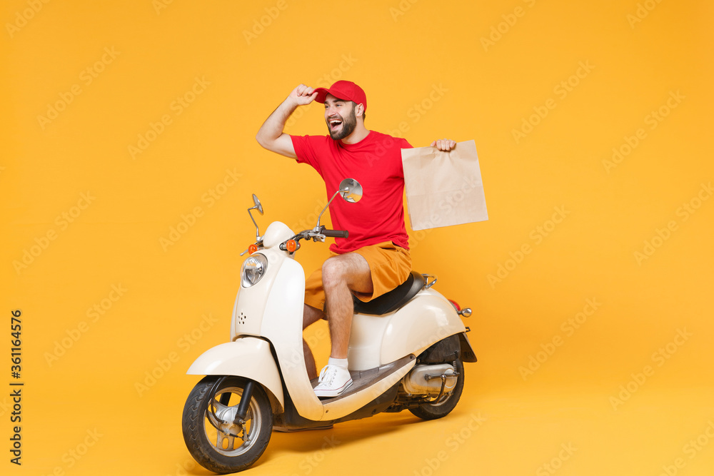 Delivery man in red cap t-shirt uniform driving moped motorbike scooter hold craft paper packet with food isolated on yellow background studio Guy employee working courier Service quarantine concept Delivery man in red cap t-shirt uniform driving moped motorbike scooter hold craft paper packet with food isolated on yellow background studio Guy employee working courier Service quarantine concept