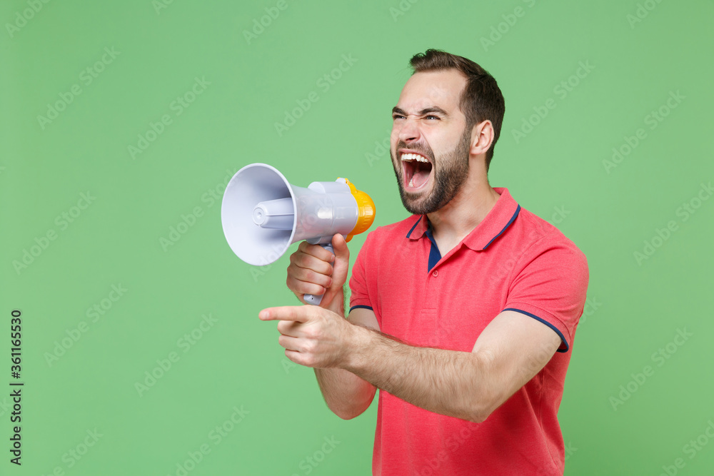 Irritated young bearded man guy in casual red pink t-shirt posing isolated on green background studio. People lifestyle concept. Mock up copy space. Screaming in megaphone pointing index finger aside. Irritated young bearded man guy in casual red pink t-shirt posing isolated on green background studio. People lifestyle concept. Mock up copy space. Screaming in megaphone pointing index finger aside.