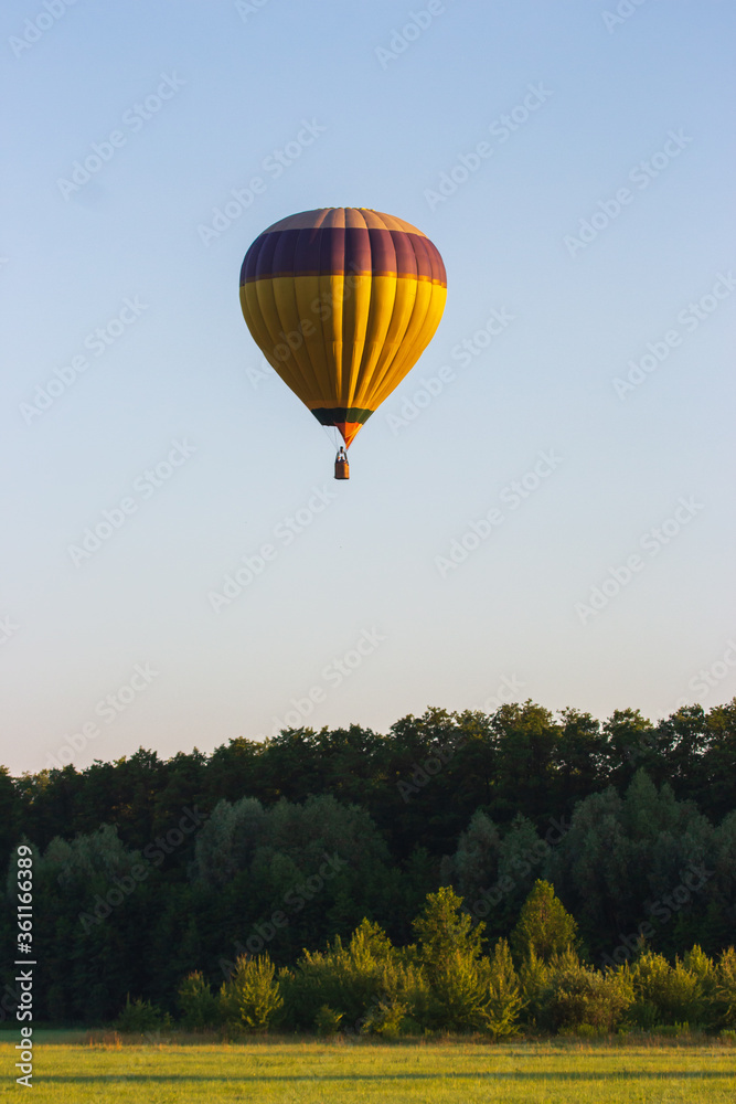 Kyiv, Ukraine - 06/26/2020: Hot air balloon in clear summer sky over trees. Yellow balloon on blue sky background. Bright geometric design. Summer leisure. Hot air balloon in flight in the morning.