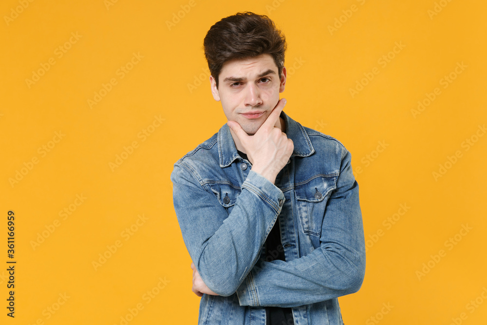 Puzzled young man guy 20s in casual denim jacket posing isolated on yellow wall background studio portrait. People sincere emotions lifestyle concept. Mock up copy space. Put hand prop up on chin. Puzzled young man guy 20s in casual denim jacket posing isolated on yellow wall background studio portrait. People sincere emotions lifestyle concept. Mock up copy space. Put hand prop up on chin.