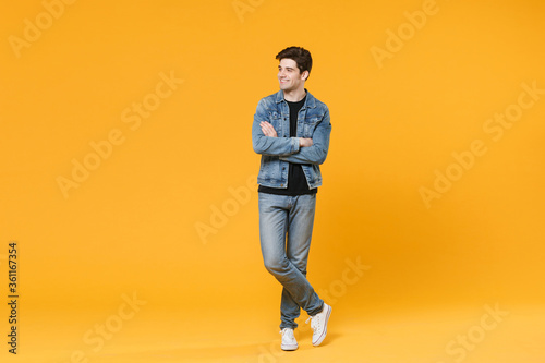 Smiling young man guy 20s wearing casual denim clothes posing isolated on yellow background studio portrait. People emotions lifestyle concept. Mock up copy space. Holding hands crossed looking aside.