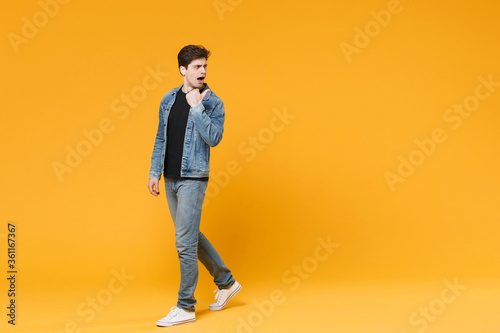 Shocked young man guy 20s wearing casual denim clothes posing isolated on yellow wall background studio portrait. People sincere emotions lifestyle concept. Mock up copy space. Pointing thumb aside.