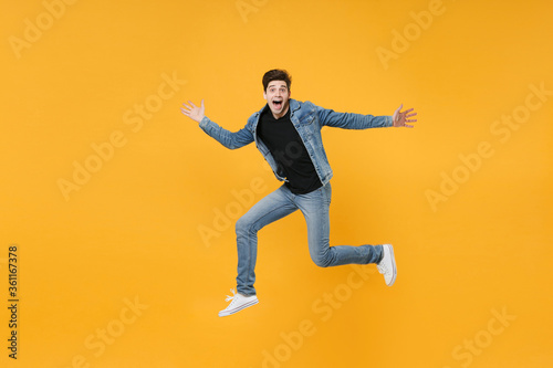 Surprised young man guy wearing casual denim clothes posing isolated on yellow background studio portrait. People sincere emotions lifestyle concept. Mock up copy space. Jumping  spreading hands legs.