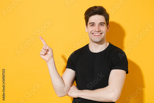 Smiling young man guy 20s in casual black t-shirt posing isolated on yellow background studio portrait. People sincere emotions lifestyle concept. Mock up copy space. Pointing index finger aside up.