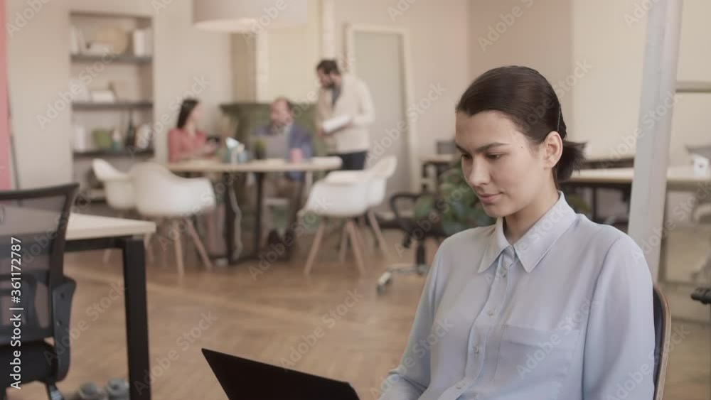Medium shot portrait of young Asian woman sitting in coworking space, using laptop then looking at camera while group of three people chatting on background Medium shot portrait of young Asian woman sitting in coworking space, using laptop then looking at camera while group of three people chatting on background