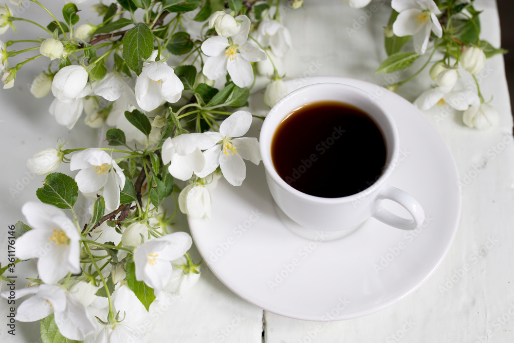 White cup of morning espresso with a blossoming apple tree branch on a wooden surface of a table with reflections. White cup of morning espresso with a blossoming apple tree branch on a wooden surface of a table with reflections.