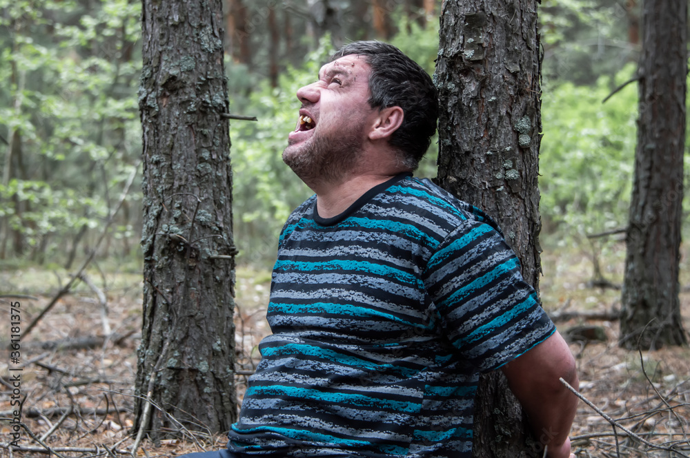 Murder in the woods. The body of a man in a blue t-shirt and trousers is sitting on the ground among the trees in the forest. Victim of an attack. Horizontal photo. Murder in the woods. The body of a man in a blue t-shirt and trousers is sitting on the ground among the trees in the forest. Victim of an attack. Horizontal photo.