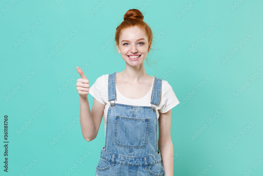 Smiling young readhead girl in casual denim clothes posing isolated on blue turquoise wall background studio portrait. People sincere emotions lifestyle concept. Mock up copy space. Showing thumb up. Smiling young readhead girl in casual denim clothes posing isolated on blue turquoise wall background studio portrait. People sincere emotions lifestyle concept. Mock up copy space. Showing thumb up.
