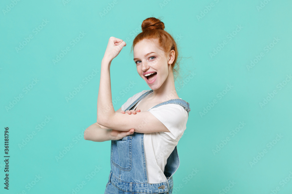 Strong young readhead girl in casual denim clothes posing isolated on blue turquoise background studio portrait. People sincere emotions lifestyle concept. Mock up copy space. Showing biceps muscles.