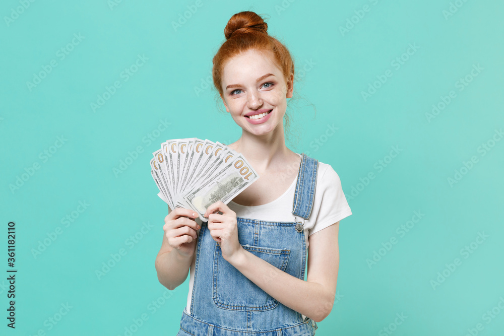 Smiling young readhead girl in casual denim clothes isolated on blue turquoise background studio portrait. People lifestyle concept. Mock up copy space. Hold fan of cash money in dollar banknotes. Smiling young readhead girl in casual denim clothes isolated on blue turquoise background studio portrait. People lifestyle concept. Mock up copy space. Hold fan of cash money in dollar banknotes.