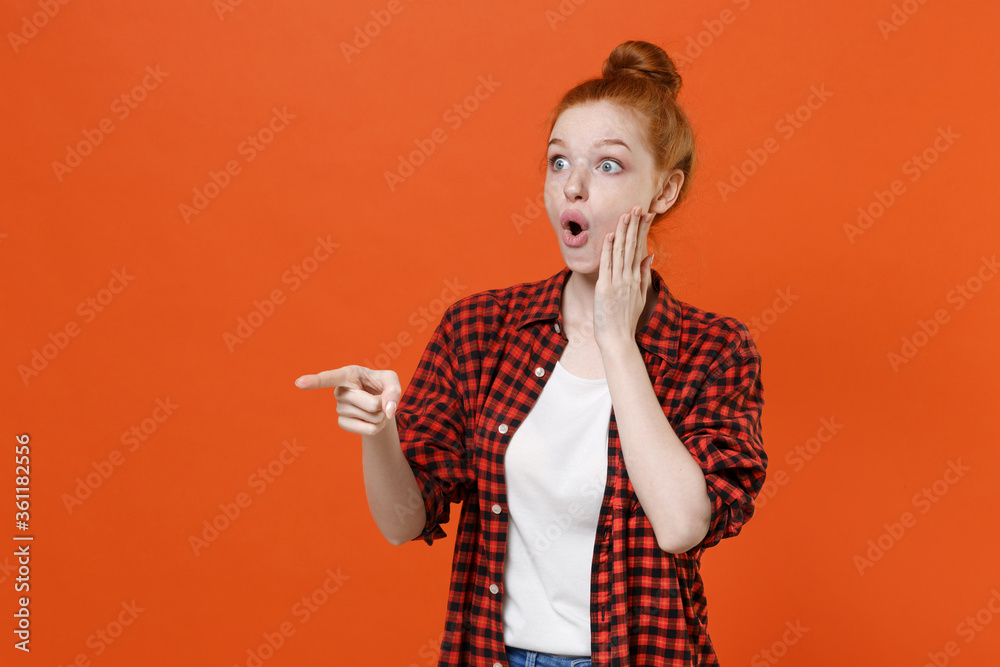 Shocked young readhead girl in casual red checkered shirt posing isolated on orange wall background studio. People lifestyle concept. Mock up copy space. Pointing index finger aside put hand on cheek. Shocked young readhead girl in casual red checkered shirt posing isolated on orange wall background studio. People lifestyle concept. Mock up copy space. Pointing index finger aside put hand on cheek.