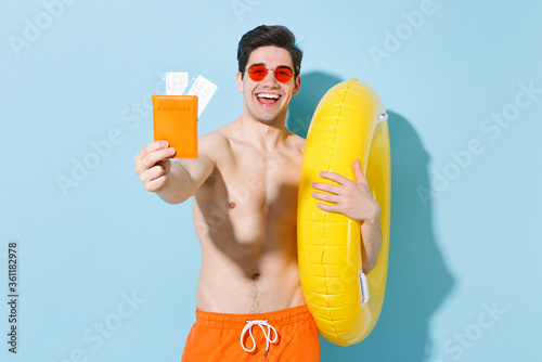 Cheerful young man guy in eyeglasses isolated on pastel blue wall background studio portrait. People summer vacation rest lifestyle concept. Mock up copy space. Hold inflatable ring passport tickets.