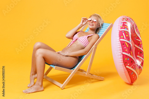 Smiling young woman in pink striped swimsuit glasses sit on deck chair isolated on yellow background studio portrait. People summer vacation rest lifestyle concept. Mock up copy space. Looking camera.