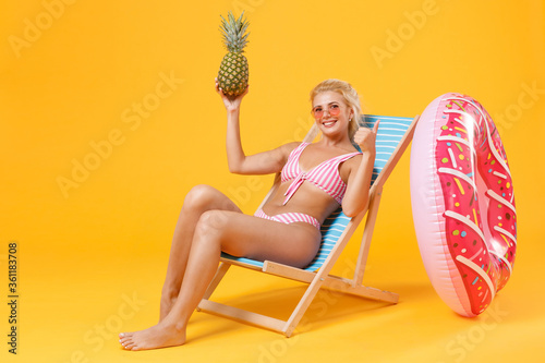 Smiling young woman girl in pink striped swimsuit glasses sit on deck chair isolated on yellow background studio. People summer vacation rest lifestyle concept. Hold pineapple fruit  showing thumb up.