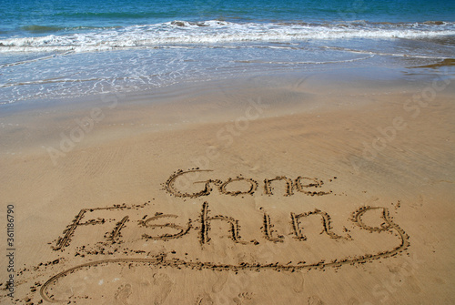 Картината върху платно Gone fishing written in sand on the beach with surf and blue sea beyond