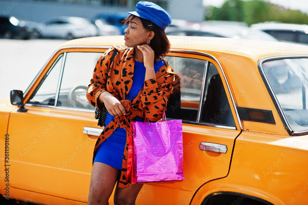 Naklejka premium Beautiful african american lady with shopping bags standing near orange classic retro car.