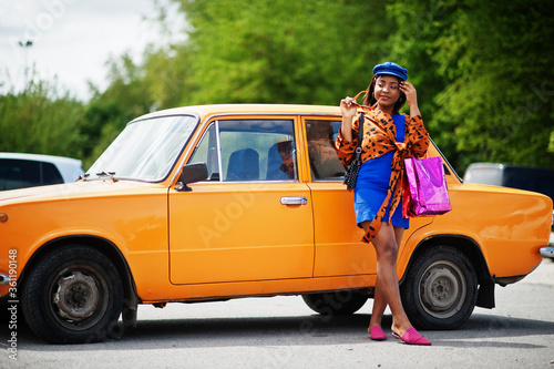 Beautiful african american lady with shopping bags standing near orange classic retro car.