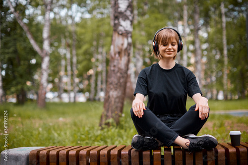 Young smiling girl in wireless headphones listening to music and meditating in lotus position in park with cup of tea