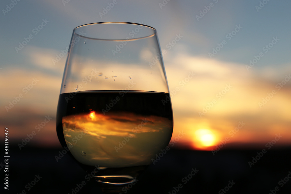 Glass with white wine on beautiful sunset background, sun and sky are reflected in alcohol drink. Concept of celebration, evening party at resort, romantic dinner outdoors