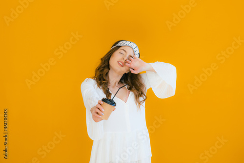 Smiling young woman wearing white dress standing isolated over yellow background, holding takeaway coffee cup