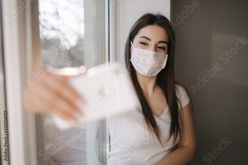 Attractive woman in medical mask make selfie at home. Female in white t'shirt and white medical mask use phone