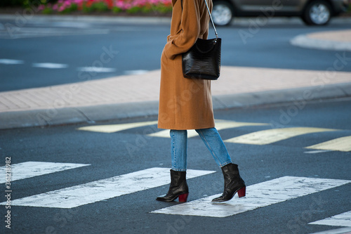 Fashionable young woman in black cowboy boots, blue jeans, beige coat, white t-shirt and crocodile print handbag in hand on the city street. Street style.