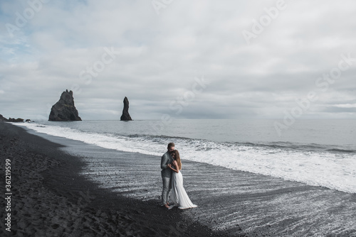 Destination Iceland wedding. A wedding couple is walking along the black beach of Vic. Sandy beach with black sand on the shores of the Atlantic Ocean. Huge foamy waves. The groom hugs the bride.