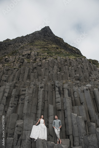 A wedding couple stands on a wall of stone pillars. The bride and groom are hugging on basalt Kekurs, on the black sandy beach of Vik. Destination Iceland wedding.