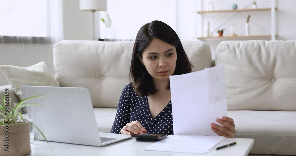 Vietnamese woman sit at coffee table in living room holding paper checking domestic bills calculates data feels upset due high prices or debt. Lack of finances, financial problems, bankruptcy concept Vietnamese woman sit at coffee table in living room holding paper checking domestic bills calculates data feels upset due high prices or debt. Lack of finances, financial problems, bankruptcy concept