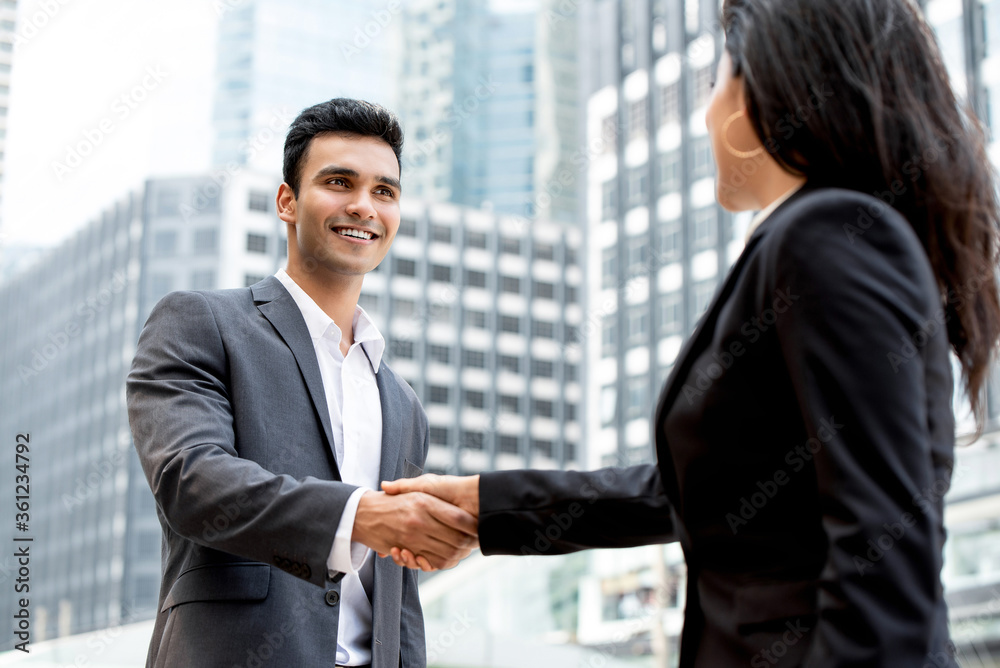 Smiling handsome young Indian businessman making handshake with businesswoman in the city with office buildings in background Smiling handsome young Indian businessman making handshake with businesswoman in the city with office buildings in background