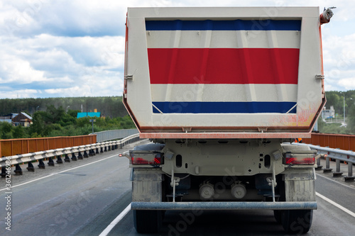 Big dirty truck with the national flag of Costa Rica moving on the highway, against the background of the village and forest landscape. 