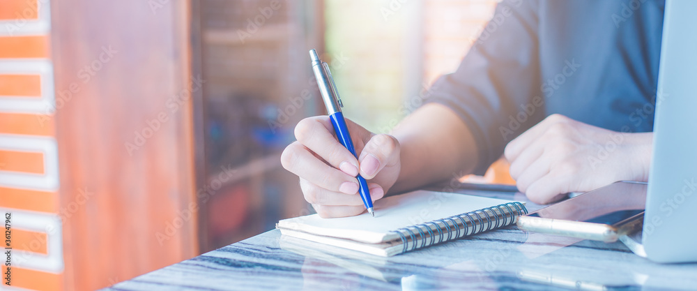 Business woman is writing on a notebook with a pen and using a laptop to work in the office.