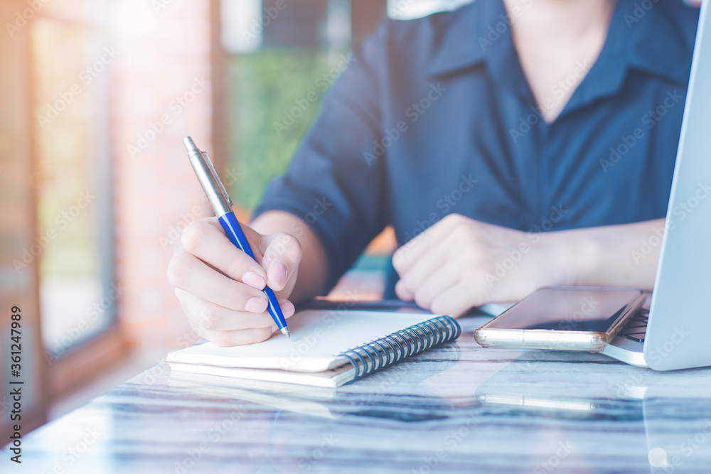 Business woman is writing on a notebook with a pen and using a laptop to work in the office.