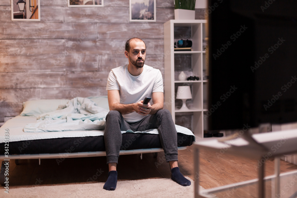 Man sitting on the edge of his bed at night watching tv holding remote control.