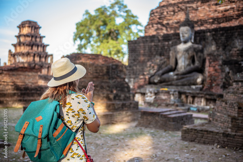 Traveler woman with backpack on ancient sculpture of a Buddha image in historical park of Sukhothai temple, Thailand.