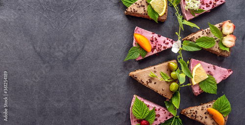 Appetizing pieces of raw mousse cakes, decorated with fruits, berries, flowers and green leaves. Black background. Concept of healthy and delicious trendy desserts