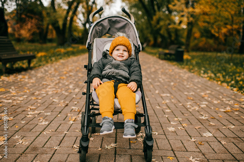 Pretty caucasian boy in yellow and grey clothes sits in a child pram in the autumn forest