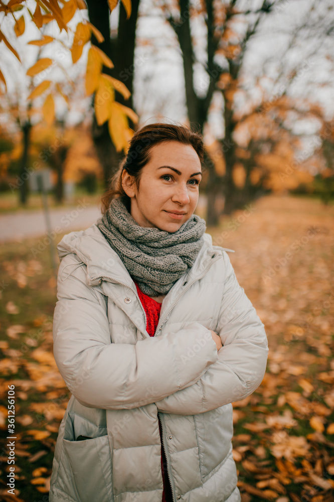 Portrait of charming caucasian female in a white warm jacket, red sweater, black pants swabbed a scarf around her neck to keep warm goes for a walk in a fall