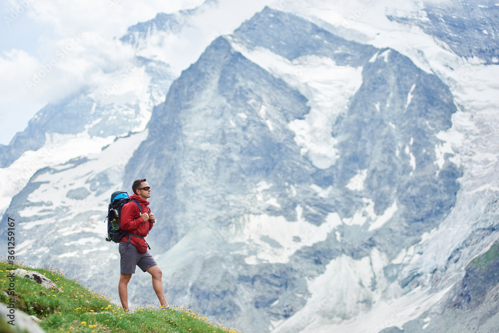 Naklejka premium Horizontal snapshot of a huge snowy mountain in Swiss Alps which takes all the background, male hiker with a backpack standing on green meadow on foreground. Concept of tourism, hiking, trekking.