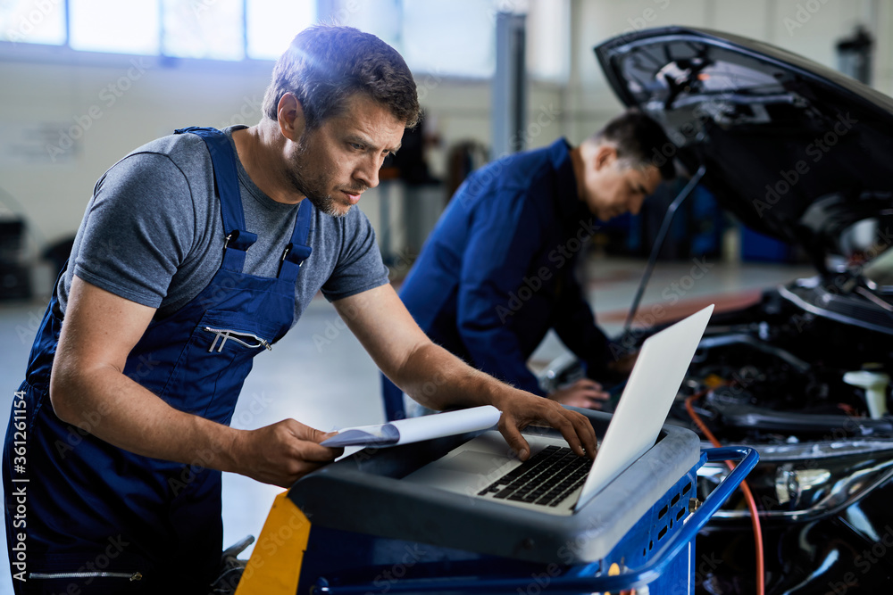 Auto repairman using laptop while working with a colleague in a workshop.
